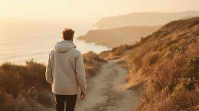 A man in a light jacket walking a coastal trail at sunrise, sea haze softening the cliffs ahead