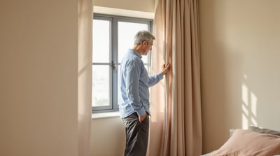 A man in blue pajamas stands beside a bare bedroom window at dusk