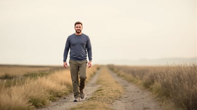 A man in a charcoal long-sleeve top and olive trousers walks toward the camera along a dirt path through open grassland