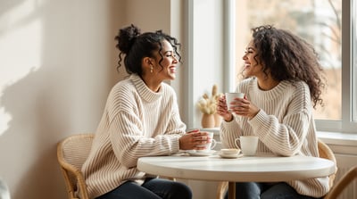 Two friends in soft knit sweaters laughing together over coffee at a sunlit cafe table by a window