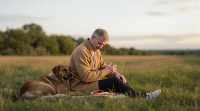 A man sits on the grass beside a dog in a meadow holding a dog lead