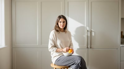 A woman sits on a wooden stool at a kitchen counter, peeling an orange over a plain bowl