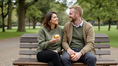 Two friends sit together on a park bench under trees holding an apple