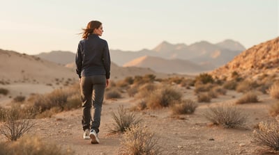A woman in a navy jacket and charcoal trousers pauses on a quiet desert trail at dawn