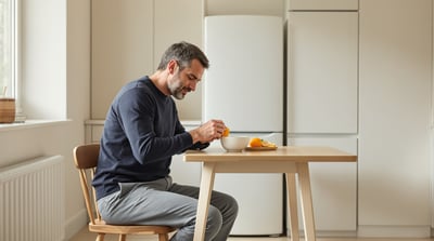 A man in a navy crew-neck jumper and full-length grey trousers sits at a kitchen table peeling an orange
