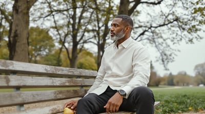 A man sits on a park bench under trees holding a pear